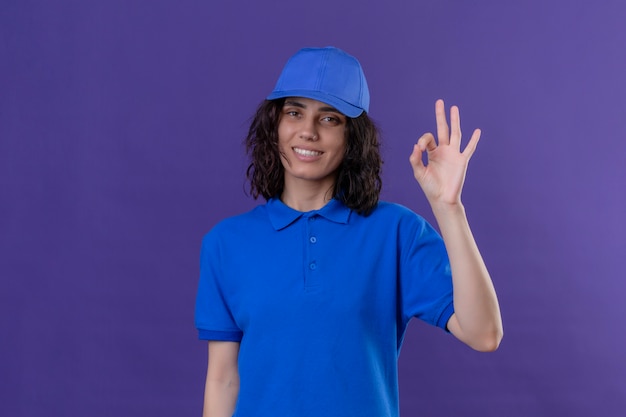 Fille de livraison en uniforme bleu et cap à joyeux sourire joyeusement faisant signe ok debout sur violet