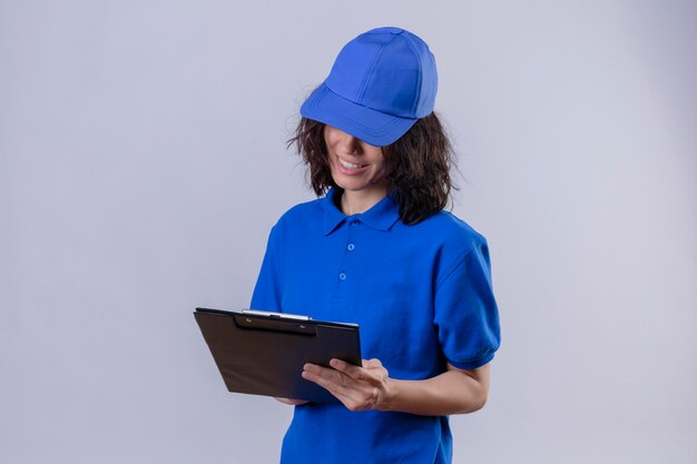 Fille de livraison en uniforme bleu et casquette tenant le presse-papiers en le regardant sourire confiant debout sur blanc