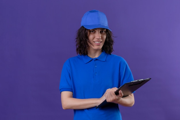 Photo gratuite fille de livraison en uniforme bleu et casquette tenant le presse-papiers avec un sourire amical debout sur violet isolé