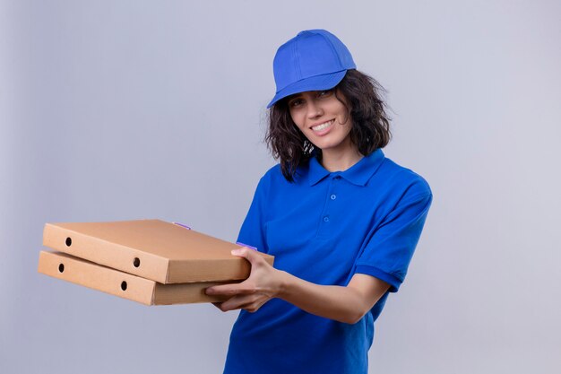 Fille de livraison en uniforme bleu et chapeau tenant des boîtes de pizza à la joyeuse positive et heureuse souriant joyeusement debout sur blanc isolé