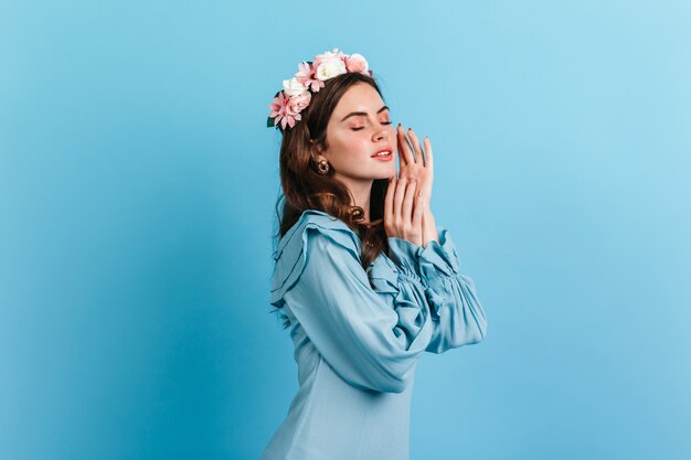 Fille avec un maquillage nude touche doucement son visage. Plan d'une brune bouclée en robe bleu ciel sur un mur isolé.