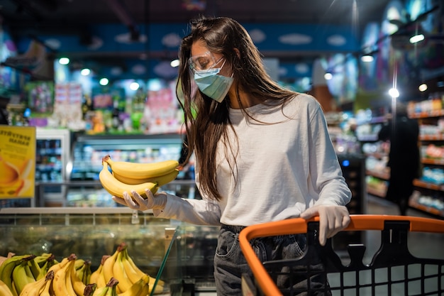 La fille avec un masque chirurgical va acheter des bananes.