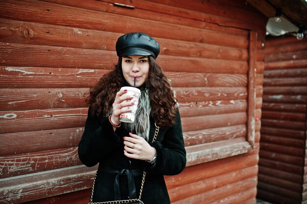 Fille mexicaine bouclée en casquette en cuir et tasse de café en plastique à portée de main contre le marché en bois