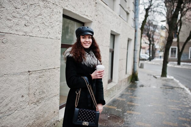 Fille mexicaine bouclée en casquette en cuir et tasse de café en plastique à portée de main marchant dans les rues de la ville