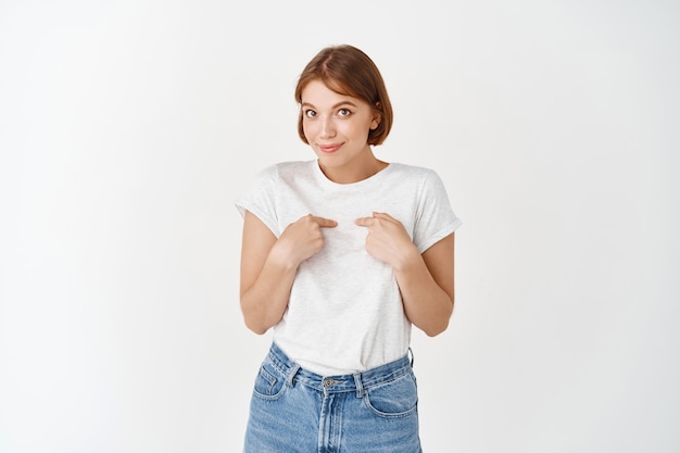 Fille mignonne timide pointant sur elle-même, mon geste, rougissant et l'air idiot, debout en t-shirt blanc et jeans sur mur blanc