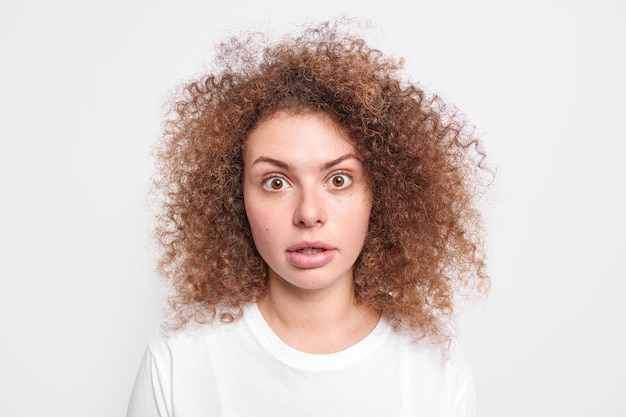 Une fille millénaire choquée aux cheveux bouclés et touffus a oublié quelque chose frappé d'une révélation surprenante, les yeux sont sortis vêtus d'un t-shirt décontracté isolé sur un mur blanc. Notion de réaction