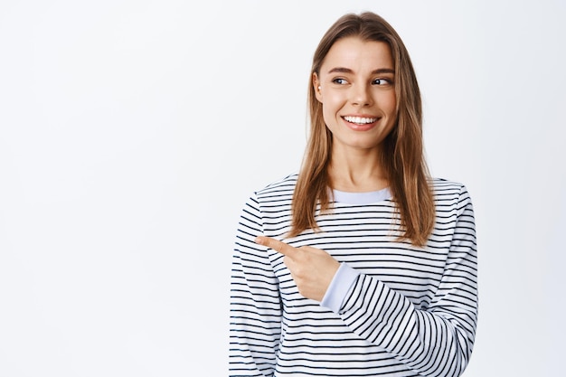 Fille millénaire élégante aux cheveux blonds et au sourire blanc parfait, pointant et regardant à gauche le logo ou la publicité, debout sur blanc