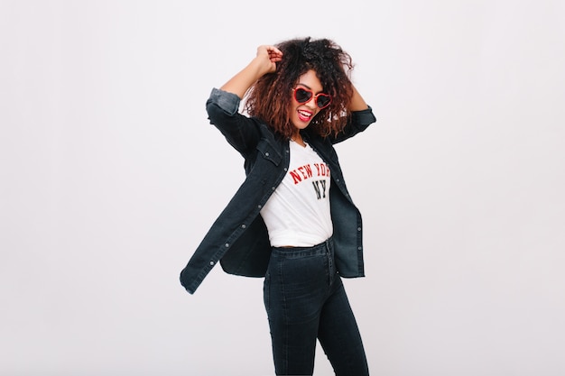 Fille mince avec une coiffure frisée en riant en dansant. Charmante jeune femme bronzée en lunettes de soleil et veste en jean s'amusant.