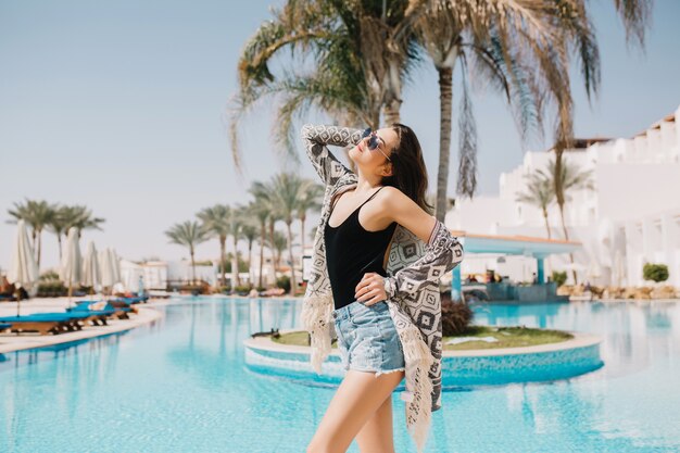 Fille mince en débardeur noir posant gracieusement près de la piscine en plein air avec des palmiers. Belle jeune femme aux cheveux raides foncés reposant à l'extérieur en vacances d'été sur station exotique