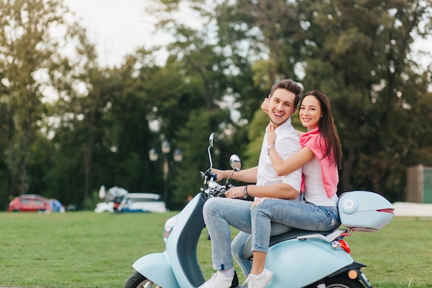 Fille mince en tenue d'été tenant les épaules de l'homme, roulant avec lui en scooter. Portrait en plein air d'un modèle masculin caucasien riant assis sur un véhicule sur fond de forêt.