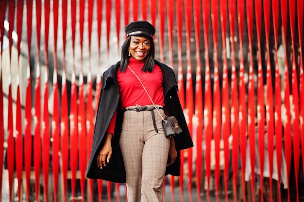 Fille de mode afro-américaine en manteau et casquette de gavroche posée dans la rue sur fond rouge