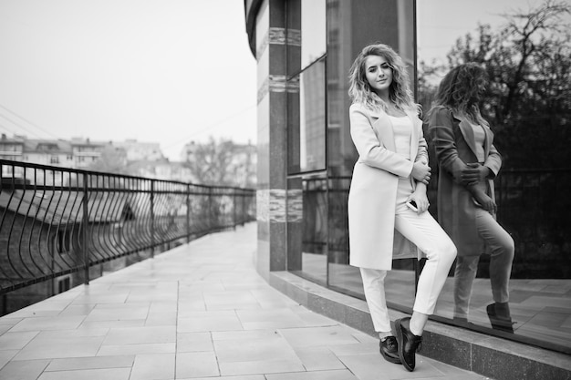 Fille modèle blonde bouclée élégante porter sur blanc avec téléphone portable à portée de main posant contre une grande fenêtre