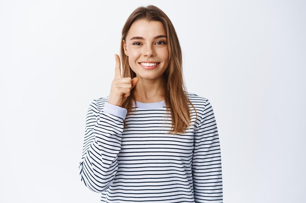 Fille moderne et élégante aux cheveux blonds et perçants, pointant le doigt vers le haut et souriant, ayant une idée ou une suggestion, portant des vêtements décontractés, blanc