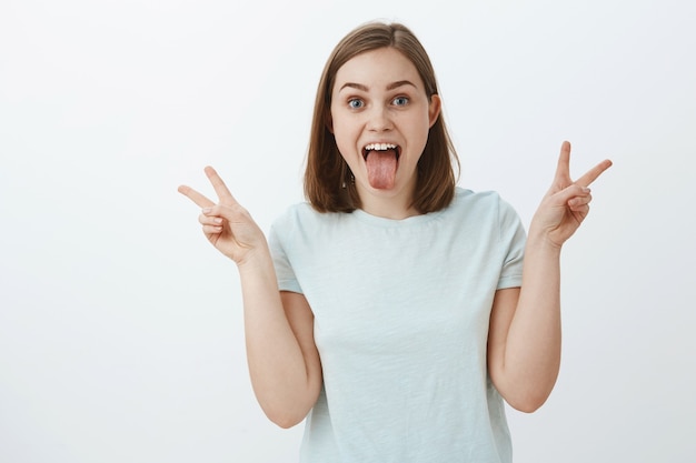 Fille n'a pas peur de s'exprimer. Joyeuse et ludique heureuse femme confiante en t-shirt décontracté montrant la langue, les signes de paix avec les deux mains s'amuser et s'amuser en profitant de la jeunesse sur le mur gris