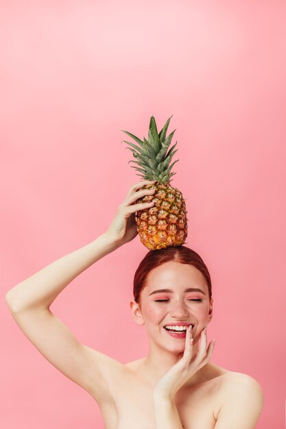 Fille nue à l'ananas souriant avec les yeux fermés. Photo de Studio de jeune femme au gingembre tenant des fruits exotiques.