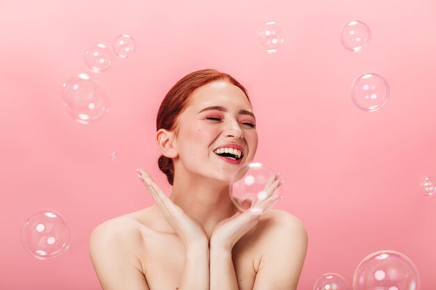 Fille nue inspirée posant avec des bulles de savon. Photo de Studio de jeune femme riante aux cheveux roux.