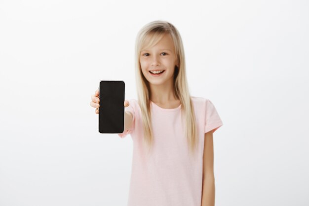 Fille optimiste montrant un nouveau téléphone mobile à des amis. Heureux enfant mignon aux cheveux blonds, tirant la main avec smartphone