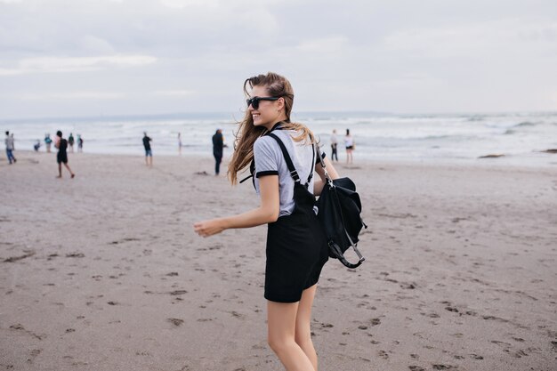 Fille pâle mince avec sac à dos noir se promenant sur la plage avec un sourire heureux. Plan extérieur d'une magnifique dame s'amusant près de la mer par temps froid.