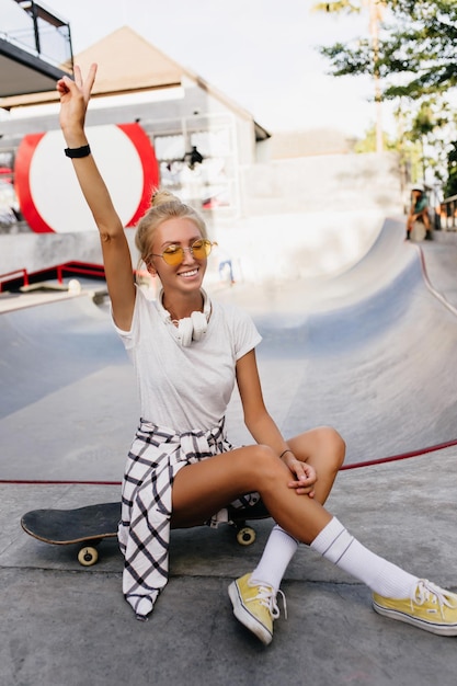 Fille de patineur bronzée gracieuse agitant la main pendant la séance photo Cheerful blonde woman in white tshirt sitting on skateboard