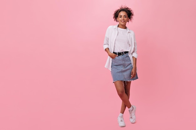 Fille à la peau foncée en jupe en jean et t-shirt blanc posant sur fond rose Jolie femme en tee-shirt souriant sur fond isolé