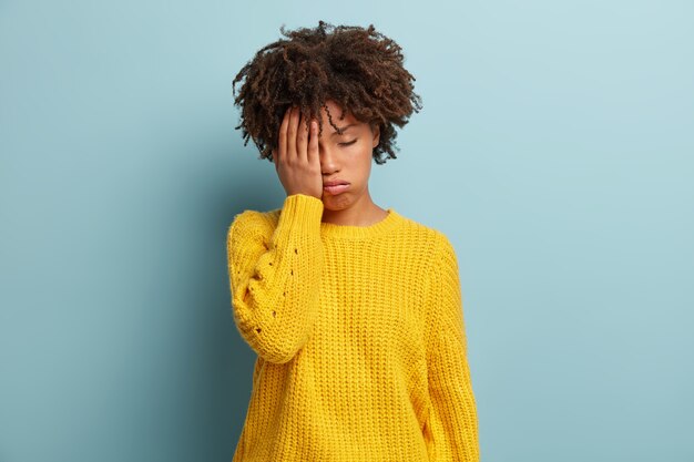 Une fille à la peau sombre surmenée et fatiguée a une expression endormie, un regard sombre, se couvre le visage avec la main, a les yeux fermés, halète de fatigue, porte des modèles de vêtements jaunes sur un mur bleu, fatigue après la fête