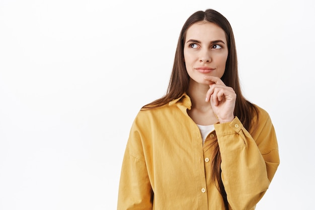 Fille pensive prenant une décision, touchant le menton et regardant de côté un choix réfléchi, méditant, debout contre un mur blanc et inventant une idée, cherchant une solution