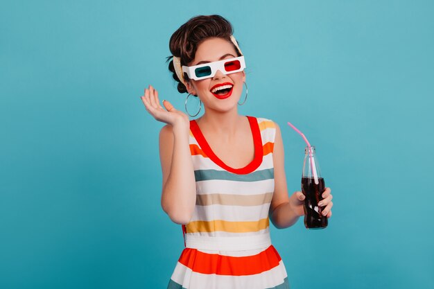 Fille de pin-up de bonne humeur posant dans des lunettes 3D. Photo de Studio de femme brune heureuse avec une bouteille de soda isolé sur fond bleu.