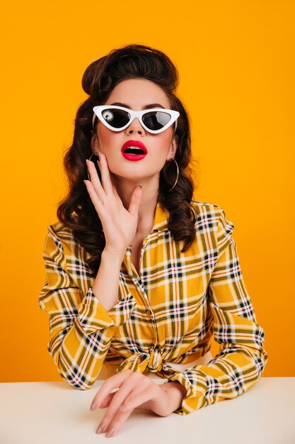 Fille de pin-up intéressée posant dans des lunettes de soleil élégantes. Photo de Studio d'une charmante dame aux cheveux bruns en chemise à carreaux.