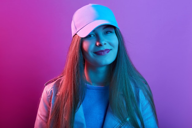 Fille portant casquette et veste en cuir regardant la caméra avec un sourire charmant, a une pommade rose, debout contre le néon rose