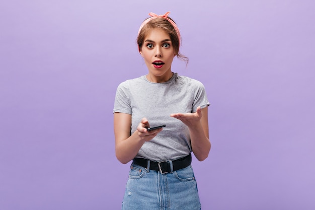 Fille pose avec malentendu et tient le téléphone. Femme cool en bandana rose avec rouge à lèvres rouge à la recherche dans la caméra sur fond isolé.