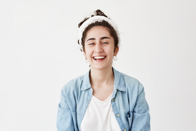 Une fille positive aux cheveux noirs porte un chiffon et une chemise en jean, a l'air heureuse, sourit largement, rit de la blague, réjouit les jeunes. Modèle féminin pose contre le mur blanc