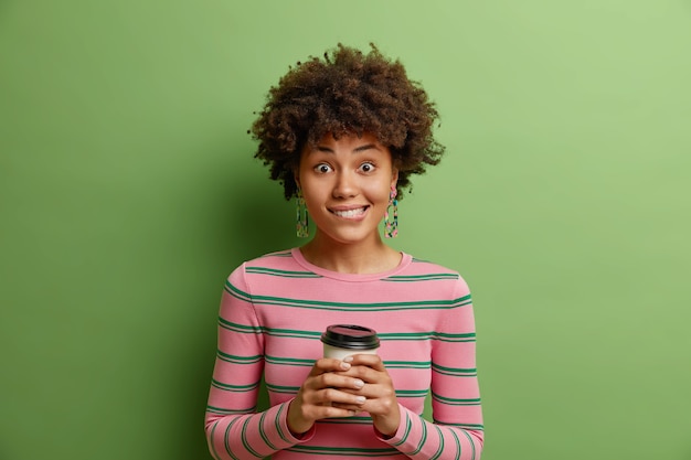 Une fille positive tient une tasse de café mord les lèvres regarde avec bonheur la caméra a une conversation agréable avec un interlocuteur porte un pull rayé et des boucles d'oreilles isolés sur un mur vert