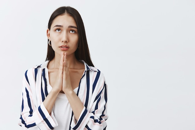 Photo gratuite fille priant dieu, demandant miséricorde et chance. portrait de jolie jeune femme concentrée en chemisier rayé, tenant la main en priant, regardant avec une expression rêveuse, espérant, être croyant