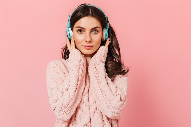 Fille en pull tricoté appuie sur les écouteurs pour un meilleur son. Femme aux yeux bleus avec un léger sourire se penche sur la caméra sur fond rose.