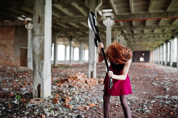 Fille punk aux cheveux rouges portant une jupe noire et rouge avec une guitare basse à un endroit abandonné Portrait d'une musicienne gothique