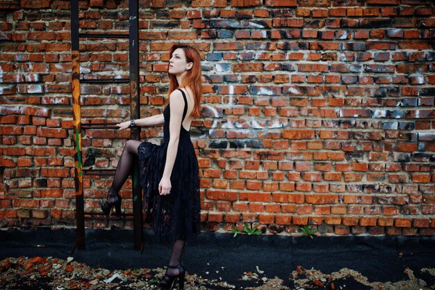 Photo gratuite fille punk aux cheveux rouges portant une robe noire sur le toit contre un mur de briques avec une échelle de fer