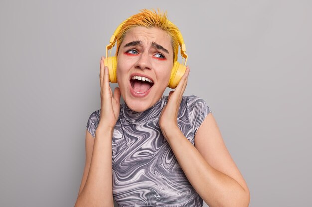 Une fille punk à la mode avec un maquillage vif et lumineux, une coiffure jaune à la mode chante une chanson apprécie la musique populaire dans des écouteurs sans fil porte un t-shirt décontracté isolé sur un mur gris attrape tout