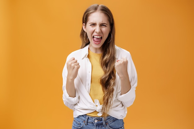 Photo gratuite une fille qui sent le pouvoir surmonte tout problème en criant pour se remonter le moral et renforcer la confiance en serrant les poings levés dans un geste joyeux et de victoire en criant à haute voix, libérant le stress sur fond orange.