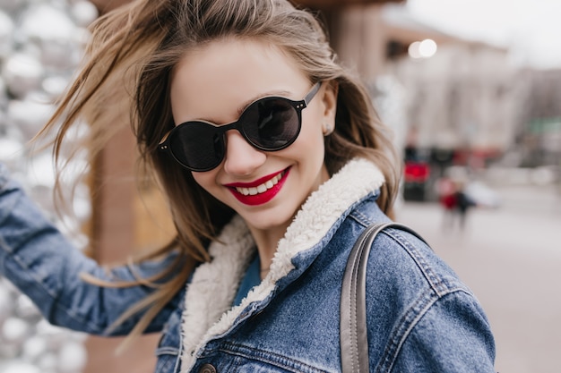 Fille de race blanche avec une expression de visage heureux se promener dans la ville au printemps matin et rire. Plan extérieur d'une magnifique femme blanche portant des lunettes de soleil sombres et portant une tenue en denim.