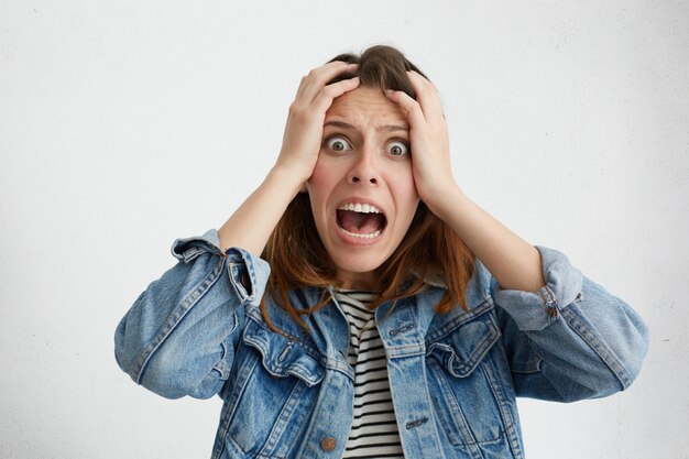 Fille de race blanche panique choquée vêtue de vêtements élégants tenant les mains sur la tête et criant de désespoir et de frustration