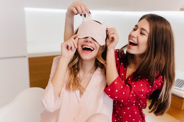 Fille raffinée en tenue rose porte un masque de sommeil le matin en riant dans la cuisine. Photo intérieure d'une jolie femme brune en pyjama rouge s'amusant avec sa sœur.