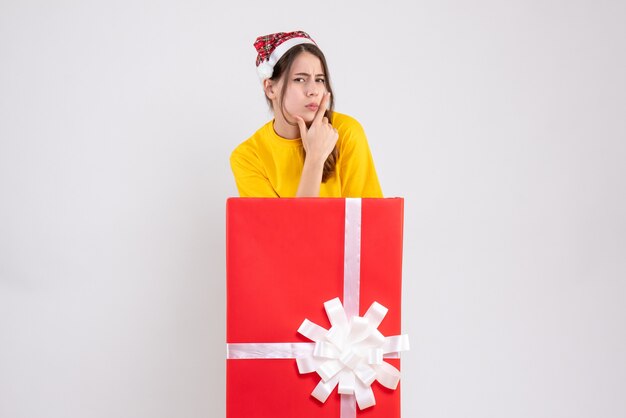 Fille réfléchie avec bonnet de Noel debout derrière un grand cadeau de Noël sur blanc