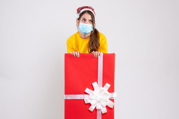 Fille réfléchie avec bonnet de Noel debout derrière un grand cadeau de Noël sur blanc