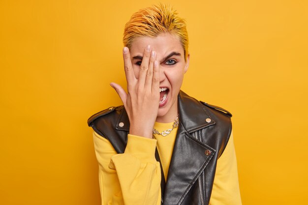 fille regarde à travers les doigts contre le visage avec la main a une coiffure à la mode maquillage lumineux vêtu d'une veste en cuir pull décontracté sur jaune s'amuse à l'intérieur