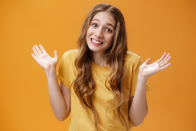 Fille rester en dehors des affaires en haussant les mains avec les mains levées et écartées, sourire stupide désolé posant sur fond orange inconscient et confus dans un t-shirt décontracté n'ayant aucune idée du sujet.