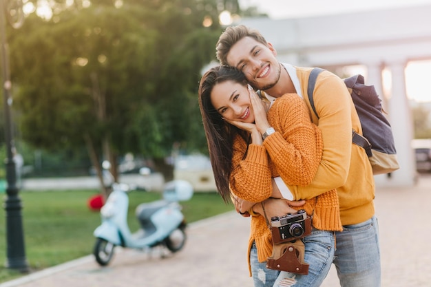 Fille De Rêve Avec Une Montre-bracelet à La Mode Riant Pendant Que Son Petit Ami La Serre Dans Ses Bras Avec Un Scooter En Arrière-plan. Heureux Jeune Homme Avec Sac à Dos Porte Une Chemise Orange S'amusant Avec Sa Petite Amie Pendant Le Repos Dans Le Parc.