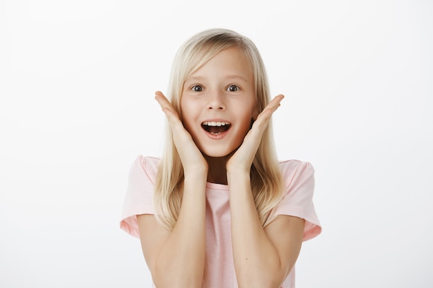 Fille rêveuse fascinée et heureuse de voir une chose incroyable. Portrait intérieur de l'enfant joyeux émotif aux cheveux blonds, souriant largement avec la bouche ouverte, tenant les paumes près des joues, admirant sur mur gris