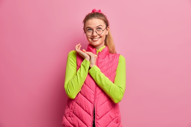 Photo gratuite fille rêveuse positive avec une expression heureuse, planifie quelque chose et se frotte les mains, concentrée au-dessus, porte des lunettes optiques, gilet rose, sourit agréablement, se tient à l'intérieur