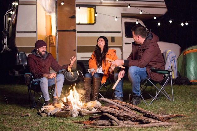 Fille riant fort après que ses amis aient raconté une blague autour d'un feu de camp avec un camping-car rétro en arrière-plan.