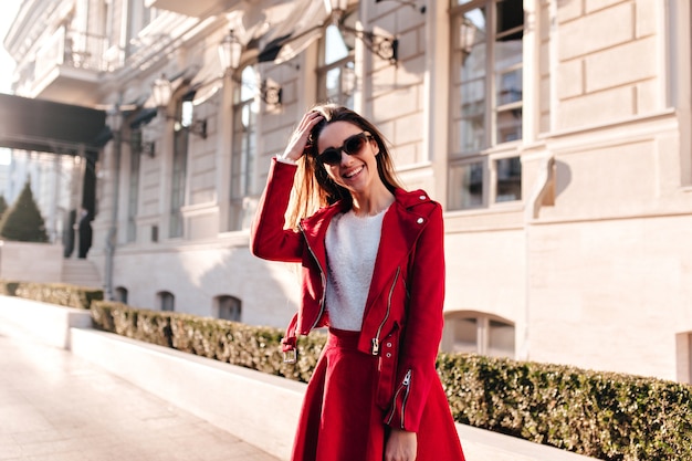 Fille riante mince dans de grandes lunettes de soleil souriant tout en posant près de l'ancien bâtiment